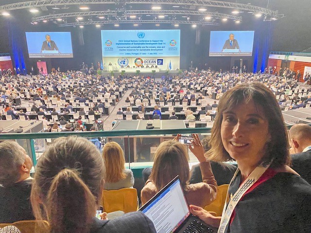 Rita sitting in the stands of the Ocean Conference Area, turning to camera with laptop open on her knees.