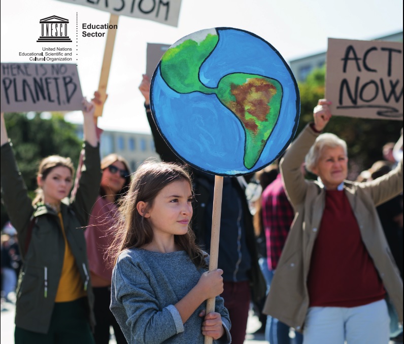 People gather together holding signs reading "Act now!" and "No Planet B". Front and centre, a girl holds a placard of a globe.