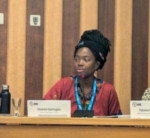 A woman sits at a conference panel table speaking into a microphone. She wears a red patterned outfit and a blue lanyard. In front of her is a nameplate that reads “WSIS – Daniella Darlington, CEO/Co‑founder Aileen AI, Ghana.” A glass, a black water bottle, and a microphone rest on the table. Wooden vertical panels form the background, and another WSIS nameplate is partially visible to her right.