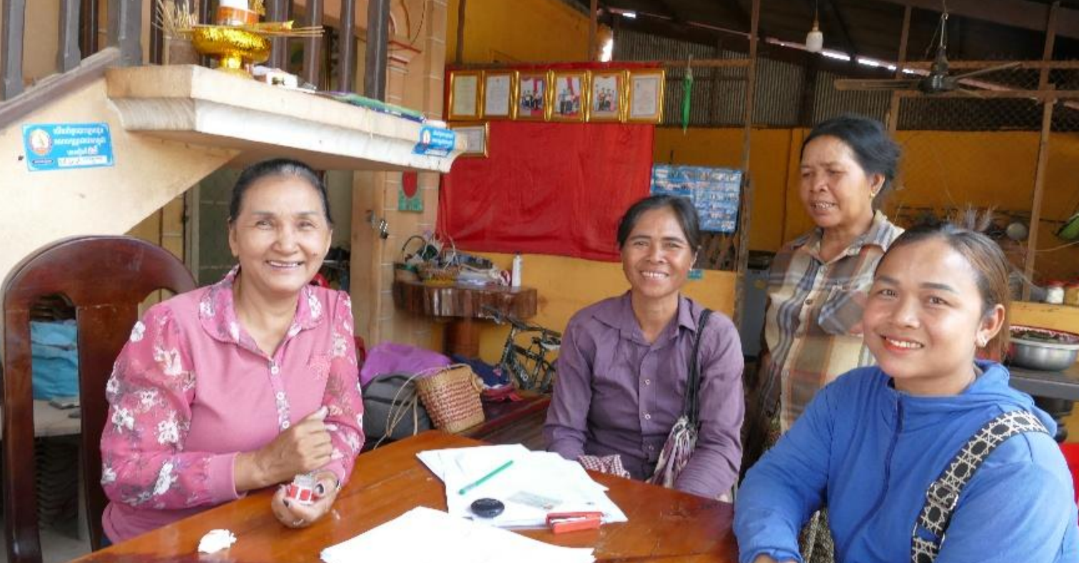 Monitoring and Evaluation Four adults sit around a wooden table in a modest indoor setting, reviewing papers and small items laid out in front of them. The room has framed pictures on a wall with a red cloth backdrop, and household items such as baskets and bowls are visible. The group appears engaged in discussion or a small meeting